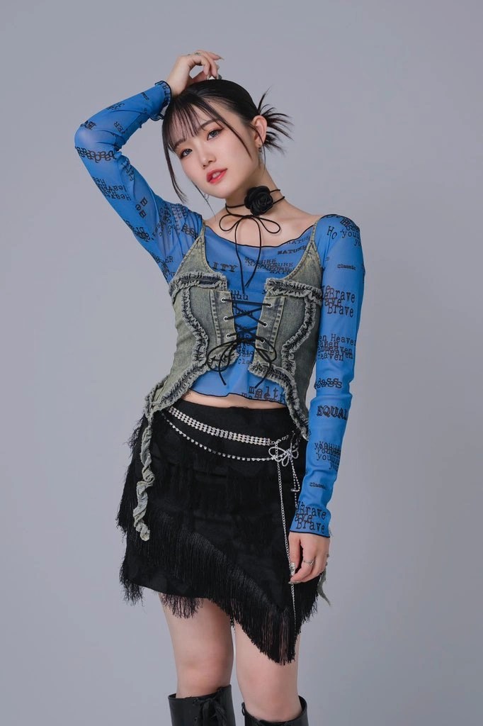 A young woman posing confidently, wearing a stylish layered outfit consisting of a blue graphic long-sleeve top, a cropped denim vest, and a black fringed skirt. She has a choker necklace and is standing against a neutral background.